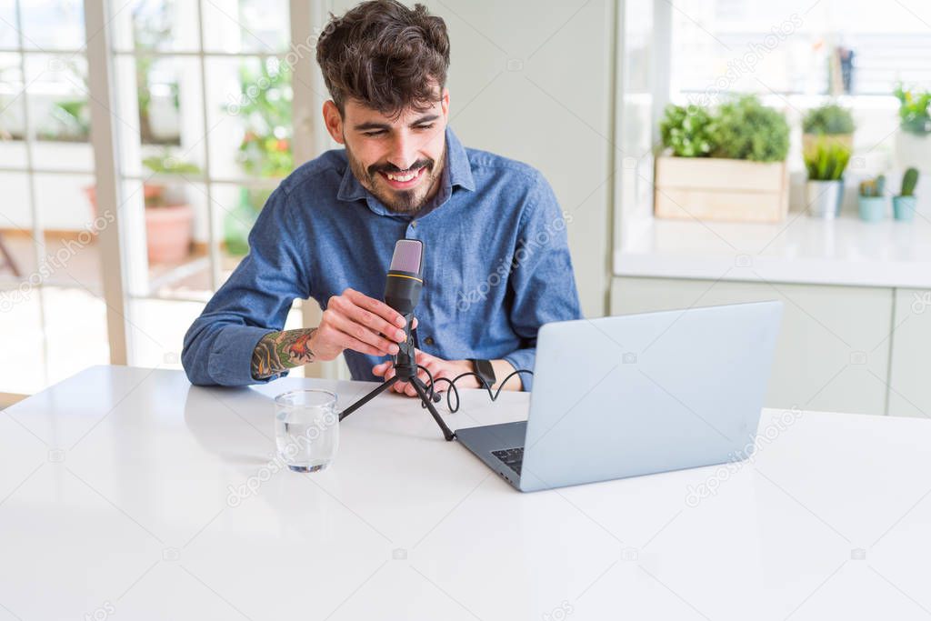 Young man recording podcast using microphone and laptop with a happy face standing and smiling with a confident smile showing teeth