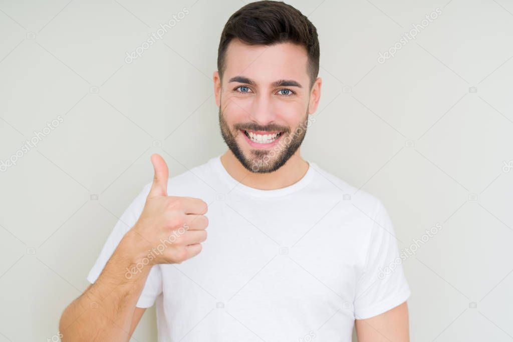 Young handsome man wearing casual white t-shirt over isolated background doing happy thumbs up gesture with hand. Approving expression looking at the camera with showing success.