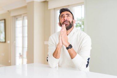 Umut ifade yüzünde çok duygusal ve endişe ile birlikte yalvarıyor ve ile dua eller casual beyaz kazak evde giyen yakışıklı İspanyol adam. Bağışlanmak. Din kavramı.