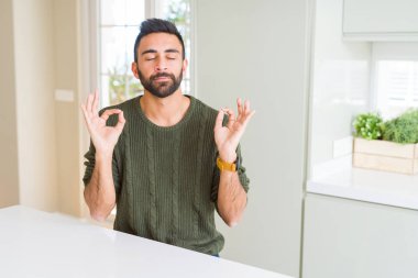 Evde rahat kazak giyen yakışıklı İspanyol adam sakin ol ve gülümseyen gözlerle kapalı parmak jestle meditasyon yapıyor. Yoga kavramı.