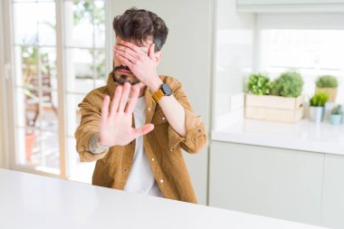 Genç adam gözleri elleriyle kapsayan beyaz masada oturan rahat ceket ve stop jest ile acı ve korku ifadesi. Utandım ve negatif kavramı.