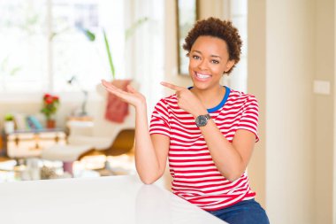 Genç güzel Afrikalı-Amerikalı kadın evde şaşırdım ve el ile sunulması ve parmak ile işaret ederken kameraya gülümseyen.