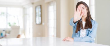 Geniş açı resim güzel genç kadının evde beyaz masaya oturan stop yapıyor şarkı elin avuç ile. Negatif ve ciddi bir jest yüz ifadesiyle uyarı.