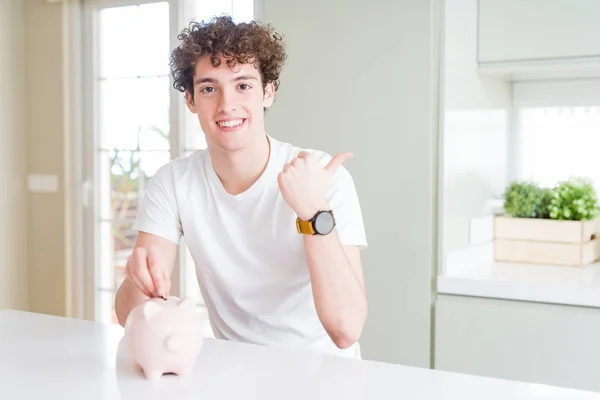 Young man investing money using piggy bank at home pointing and showing with thumb up to the side with happy face smiling