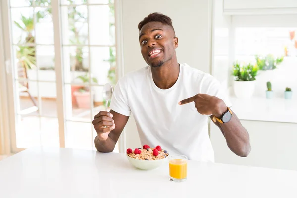 Young african american man eating healthy breakfast in the morning with surprise face pointing finger to himself
