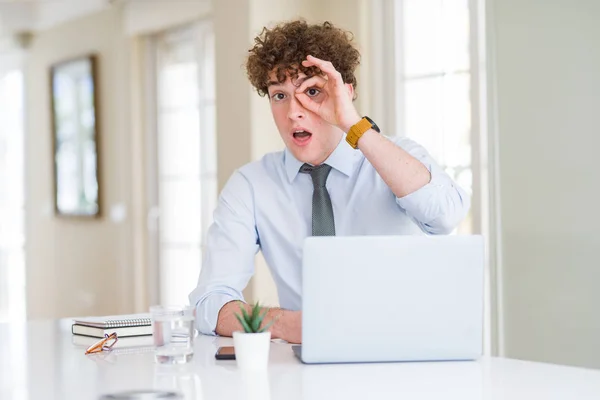Young business man working with computer laptop at the office doing ok gesture shocked with surprised face, eye looking through fingers. Unbelieving expression.