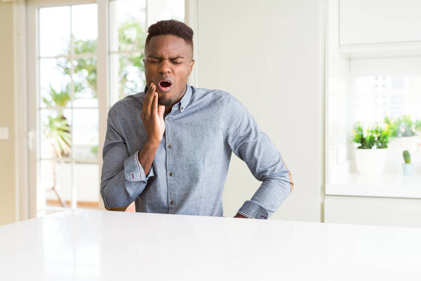 Красивый африканский американец на белом столе касается рта рукой с болезненным выражением лица из-за зубной боли или зубной болезни на зубах. Концепция дантиста
.