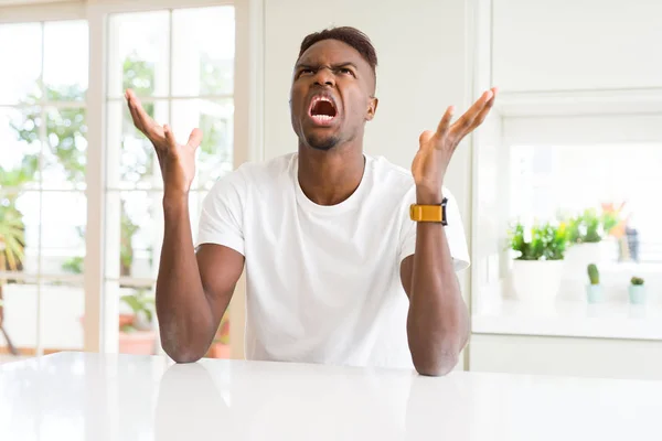 英俊的非洲裔美国男子在白色的桌子上在家里疯狂和疯狂的喊叫和大喊 咄咄逼人的表情和手臂提高 挫折概念 — 图库照片