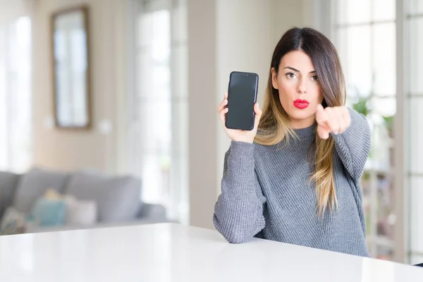 年轻美丽的女人显示智能手机屏幕在家里用手指指向相机和你 手的标志 积极和自信的手势从前面 — 图库照片
