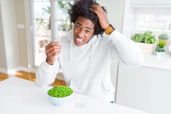 African American man eating fresh green peas at home stressed with hand on head, shocked with shame and surprise face, angry and frustrated. Fear and upset for mistake.