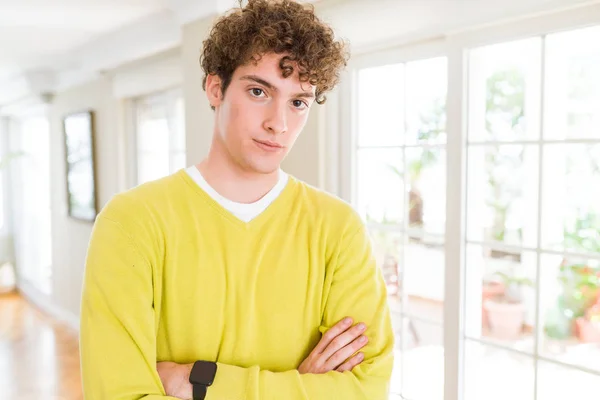 年轻英俊的男人穿着黄色毛衣在家里放松与严肃的表情在脸上 简单和自然与交叉的武器 — 图库照片