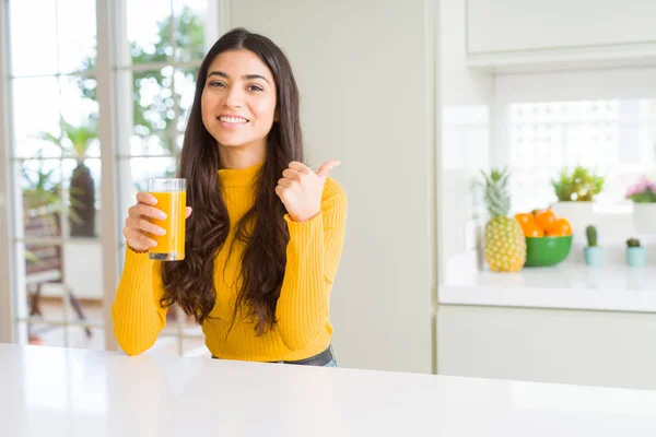 年轻女子喝了一杯新鲜的橙汁指着和愉快的脸 用拇指向一边展示 — 图库照片