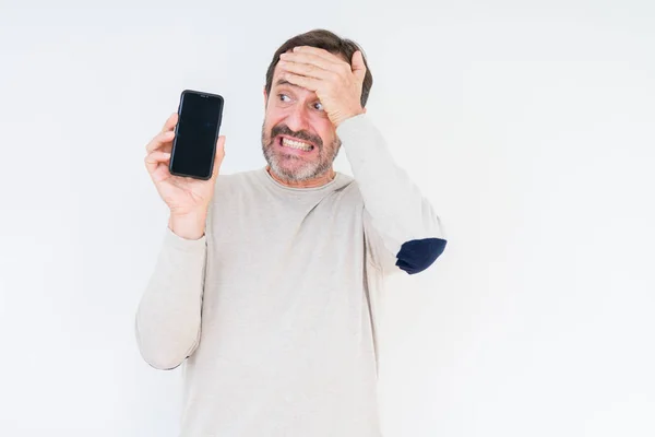 Senior man showing smartphone screen over isolated background stressed with hand on head, shocked with shame and surprise face, angry and frustrated. Fear and upset for mistake.