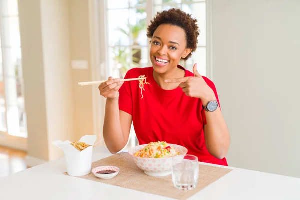 Young african american woman with afro hair eating asian food at home very happy pointing with hand and finger
