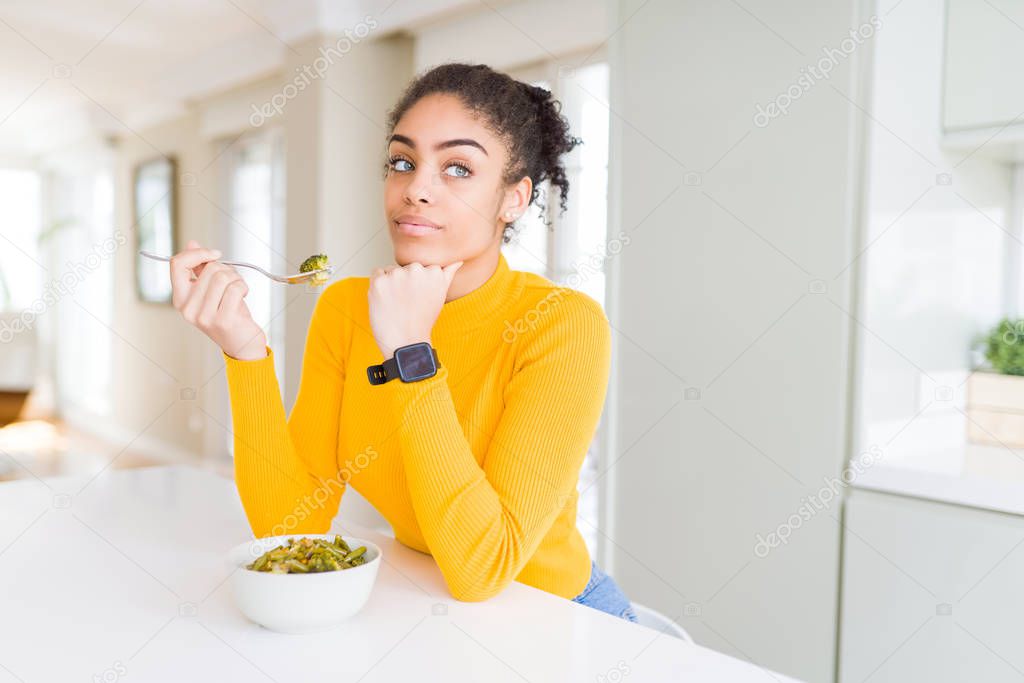 Young african american woman eating healthy green vegatables serious face thinking about question, very confused idea