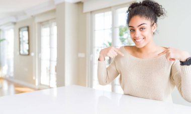 Evde masaya oturan afro saçlı güzel genç Afro-Amerikan kadın kendini gururlu ve mutlu parmakları ile işaret emin yüz, gülümseme ile seyir.