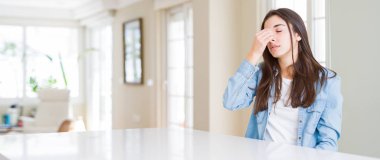 Güzel genç kadın evde beyaz masaya oturan geniş açı resim sürtünme burun ve göz yorgunluğu ve baş ağrısı duygu yorgun. Stres ve hayal kırıklığı kavramı.