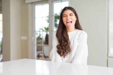 Genç güzel kadın evde beyaz masada dilini out komik ifade ile mutlu. Duygu kavramı.