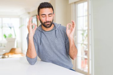 Evde rahat sweatshirt giyen yakışıklı İspanyol adam sakin ol ve gülümseyen gözlerle kapalı parmak jestle meditasyon yapıyor. Yoga kavramı.