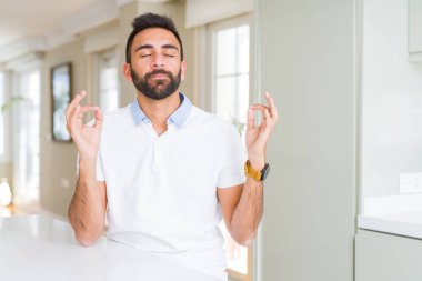 İspanyol yakışıklı casual beyaz t-shirt evde dinlenmek ve gülümseyen gözlerle parmak ile meditasyon jest yapıyor kapalı. Yoga kavramı.