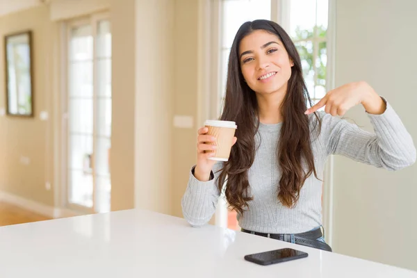 Young woman drinking a cup of coffee at home with surprise face pointing finger to himself