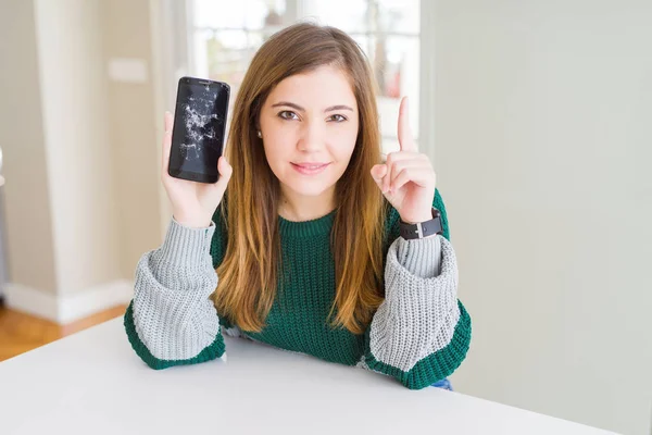 Beautiful young woman holding broken smartphone surprised with an idea or question pointing finger with happy face, number one