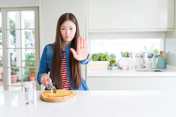 Beautiful Asian woman eating and cutting chesse pizza with open hand doing stop sign with serious and confident expression, defense gesture
