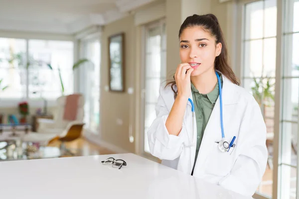 Young doctor woman wearing medical coat at the clinic looking stressed and nervous with hands on mouth biting nails. Anxiety problem.