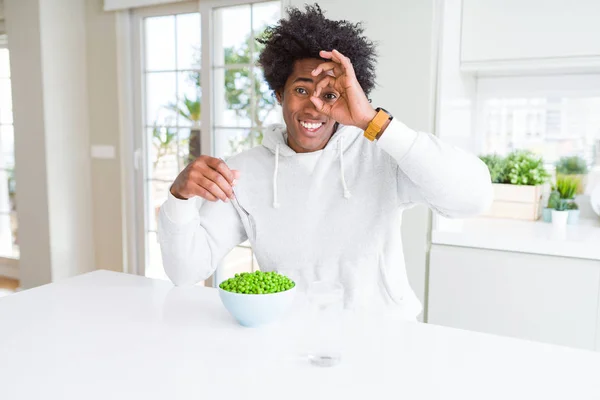 African American man eating fresh green peas at home with happy face smiling doing ok sign with hand on eye looking through fingers