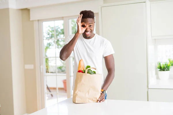African american man holding paper bag full of fresh groceries with happy face smiling doing ok sign with hand on eye looking through fingers