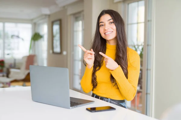 Young woman using computer laptop smiling and looking at the camera pointing with two hands and fingers to the side.