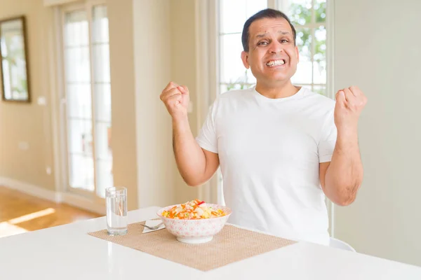 Middle age man eating rice at home celebrating surprised and amazed for success with arms raised and open eyes. Winner concept.