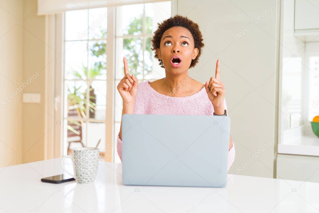Young african american woman working using computer laptop amazed and surprised looking up and pointing with fingers and raised arms.