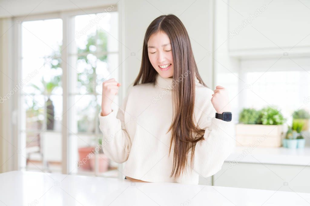 Beautiful Asian woman wearing casual sweater very happy and excited doing winner gesture with arms raised, smiling and screaming for success. Celebration concept.