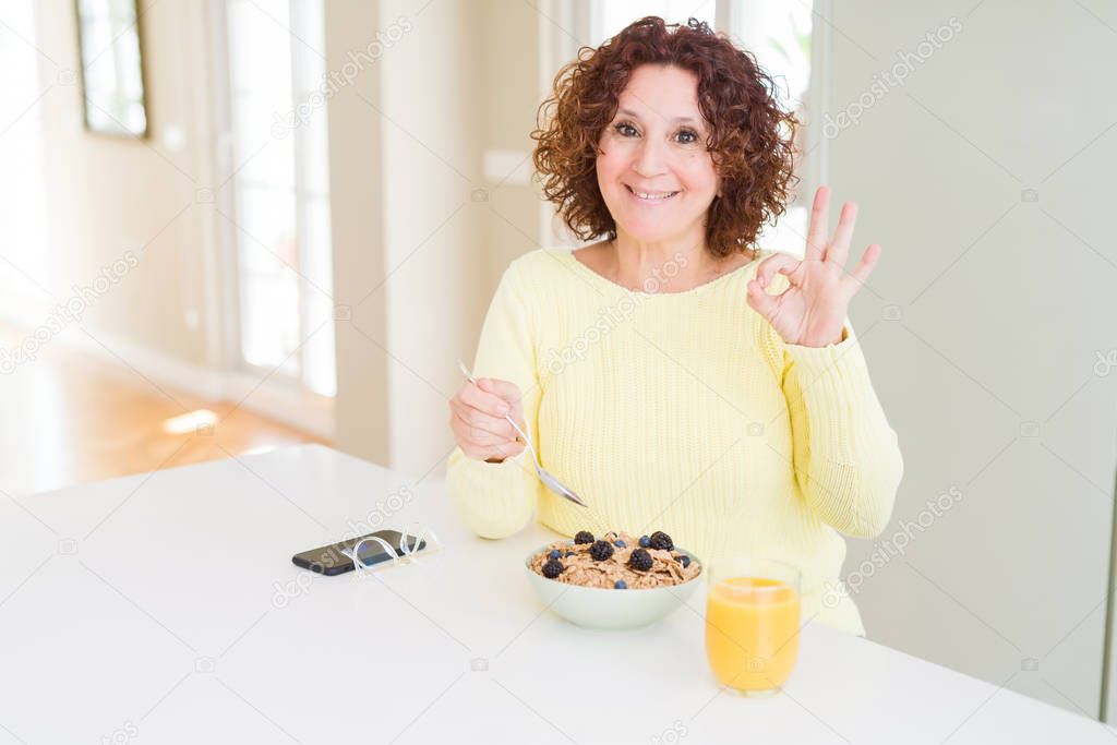 Senior woman eating healthy breakfast in the morning at home doing ok sign with fingers, excellent symbol