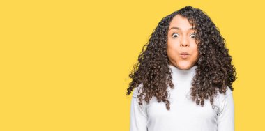 Young beautiful woman with curly hair wearing turtleneck sweater puffing cheeks with funny face. Mouth inflated with air, crazy expression. clipart