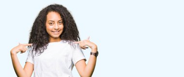 Yüzünde gülümseme ile kendinden emin görünüyorsun, kendini gururlu ve mutlu parmakları ile işaret casual beyaz t-shirt giyen kıvırcık saçlı genç güzel kız.