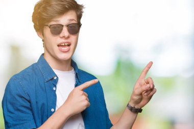 Yakışıklı delikanlı gülümseyen ve iki elleri ve parmakları ile tarafına işaret kamera bakarak izole arka plan üzerinde güneş gözlüğü takıyor.