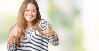 El ile pozitif jest yapıyor izole arka plan başarı işareti üzerinde çizgili Süveter giyen genç güzel esmer kadın başparmak gülümseyen ve mutlu. Neşeli bir ifadeyle, kazanan jest kameraya bakıyor.