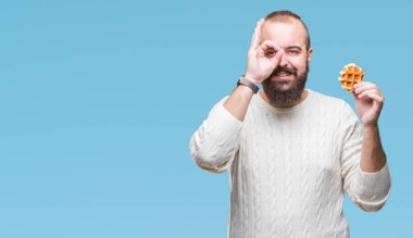 Genç beyaz hipster man ile mutlu yüz el parmaklarının arasından seyir göz üzerinde ok işaretiyle yapıyor gülümseyen izole arka plan üzerinde tatlı waffle yemek