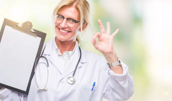 中年金发医生妇女持有剪贴板在孤立的背景下做 标志与手指 优秀的符号 — 图库照片