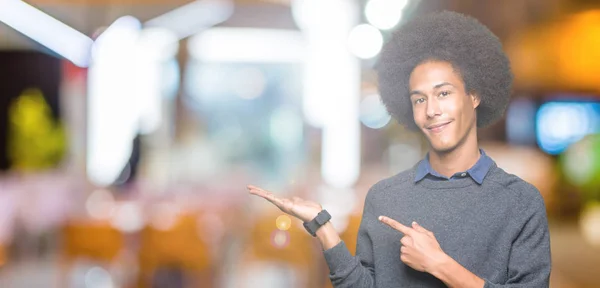 Young african american business man with afro hair amazed and smiling to the camera while presenting with hand and pointing with finger.