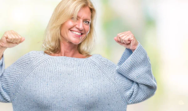 Middle age blonde woman wearing winter sweater over isolated background showing arms muscles smiling proud. Fitness concept.