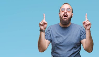 Güneş gözlüğü izole arka plan üzerinde giyen genç beyaz hipster adam hayret ve yukarıya bakıp parmakları ile işaret şaşırttı ve silah kaldırdı.