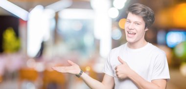 İzole arka plan gösteren palm el üzerinde rahat beyaz t-shirt giyen ve yukarıya, mutlu ve neşeli gülümseyen baş parmakları Tamam jest yapıyor yakışıklı delikanlı