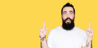 Uzun saç ve sakal casual beyaz t-shirt giyen genç hipster adamla hayrete ve yukarıya bakıyor ve parmakları ile işaret ve silah kaldırdı şaşırttı.