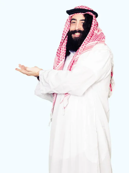 伝統的なクーフィーヤを着て長い髪をアラビアン ビジネス人間手側にポインティングをスカーフし 広告笑顔幸せと自信を持って提示 手のひらを開く — ストック写真