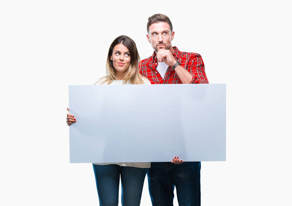 Young couple together holding blank banner over isolated background serious face thinking about question, very confused idea