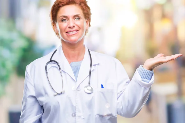 陽気な提示とカメラを見て手の手のひらで指している笑みを浮かべて分離の背景に医療制服を着て先輩の白人医師女性 — ストック写真