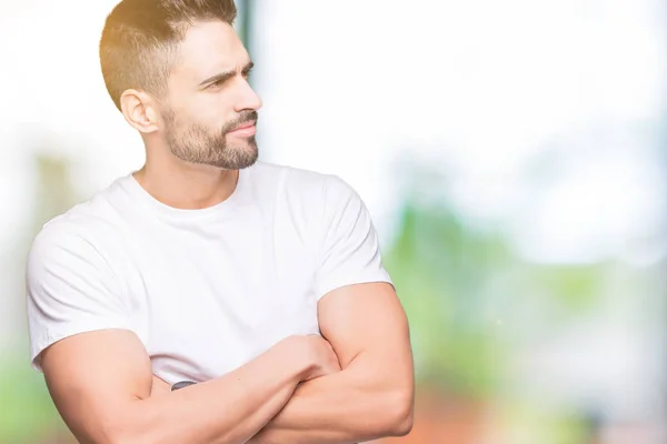 Handsome man wearing white t-shirt over outdoors background smiling looking to the side with arms crossed convinced and confident